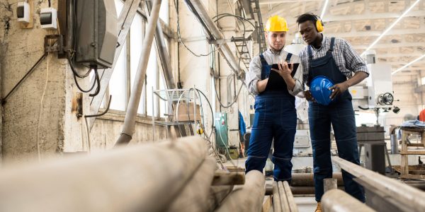 Multi-ethnic team of inspectors wearing overalls and hardhats standing at spacious production department of modern plant while making inspection, one of them writing something down